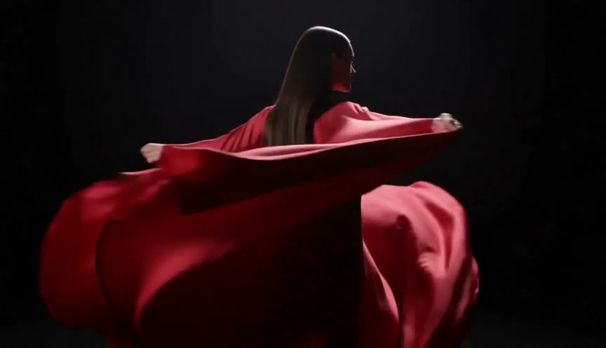 A model in a flowing scarlet gown turning as the fabric ripples in slow motion against a dark studio, with dramatic rim light and an editorial mood.